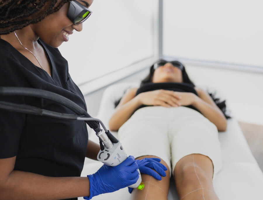 A woman receiving Laser Hair Removal treatment on her legs by a professional wearing protective eyewear and gloves in a modern clinic setting.